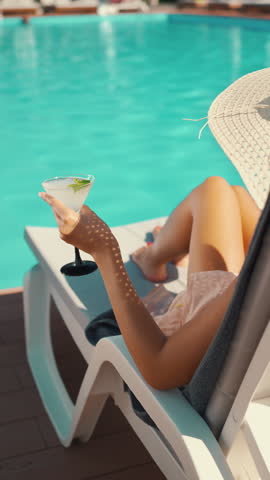 Young woman in sun hat sitting on sunbed and drinking fresh cocktail by swimming pool. Female enjoying summer vacation at luxury resort sun