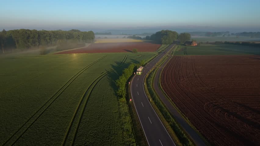 Rural two-lane highway winding through lush green crop fields and plowed farmland at golden sunrise, truck and cars in light traffic, morning mist over distant tree line and pale blue sky. Aerial view