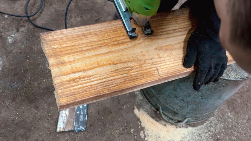 Close-up of a carpenter wearing protective gloves cutting a wooden plank with a green electric jigsaw, creating a cloud of sawdust in a workshop environment during a home improvement project