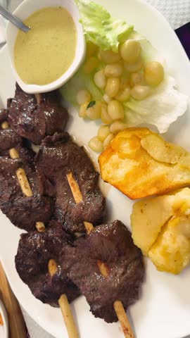 Vertical close-up of traditional Peruvian dish Anticuchos served on a plate in a restaurant. Grilled marinated beef heart skewers with boiled potatoes and spicy sauces, authentic Peruvian cuisine