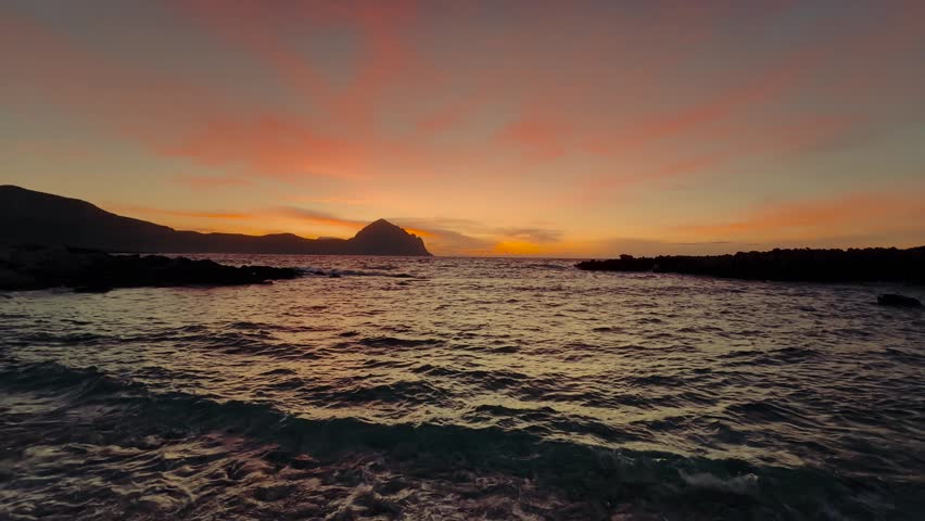 Vibrant orange sky over the sea in Sicily with the silhouette of Monte Cofano and waves. San Vito Lo Capo