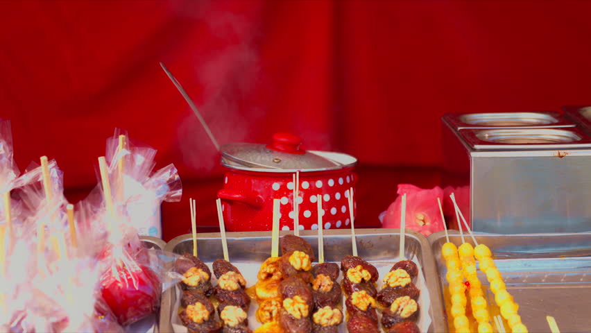 Street food stall displaying skewers, candy apples and assorted sweet snacks on metal trays, with steaming pots and wooden sticks creating an inviting market scene, 4K selective focus