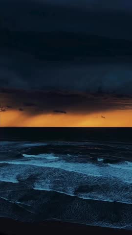Powerful waves crash against rocky shoreline under a moody, stormy sky with golden sunset hues, creating a dynamic seascape with a lone bird flying in the distance.
