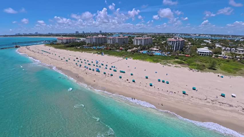 Aerial view of Singer Island beach with turquoise water, blue umbrellas on the sand, people, and luxury resorts in Riviera Beach, Florida, United States.