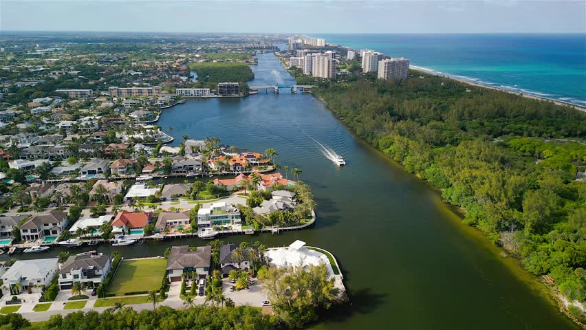 Aerial view of the Intracoastal Waterway with a boat cruising past luxury waterfront mansions and green vegetation near the Atlantic coast in Boca Raton, Florida, United States.