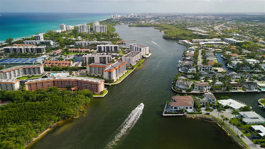 Aerial view of the Intracoastal Waterway featuring luxury residential buildings, lush greenery, and a boat cruising through the channel in Boca Raton, Florida, United States.