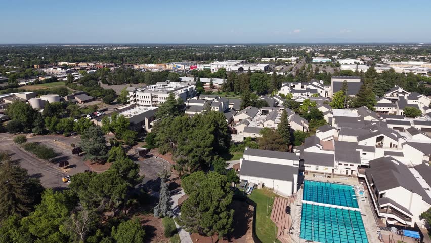 Aerial perspective of the San Joaquin Delta College campus, showcasing the swimming pool, parking lot, academic buildings, and surrounding trees on a bright, sunny day in Stockton, California