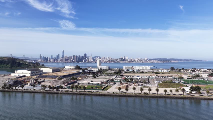 Sweeping aerial view showing the urban development on Treasure Island with the iconic San Francisco skyline and Bay Bridge visible across the water under a beautiful blue sky with wispy clouds