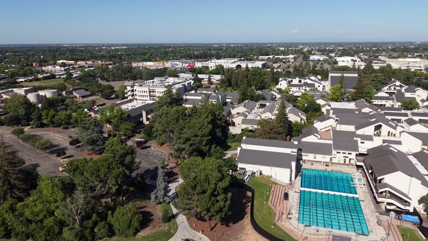 Aerial view of the San Joaquin Delta College campus in Stockton, California, showing buildings, a large swimming pool, and the surrounding cityscape under a clear blue sky on a sunny day