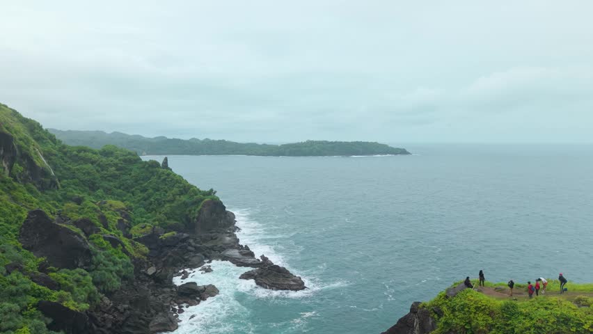 Aerial view of Bukit Pengilon featuring lush green cliffs and people standing on a ridge overlooking the ocean in Kecamatan Tepus, Daerah Istimewa Yogya, Indonesia.