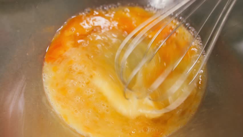 Close-up of eggs being whisked with a whisk in a transparent glass bowl. Cooking process, food preparation, baking ingredients and homemade kitchen activity concept. High quality 4k footage