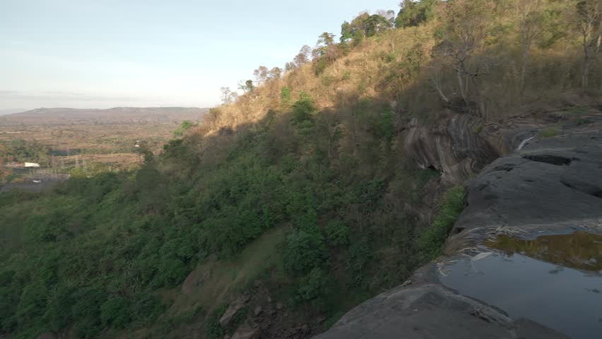 Camera tilts down over the edge of a waterfall cliff to reveal the riverbed below.