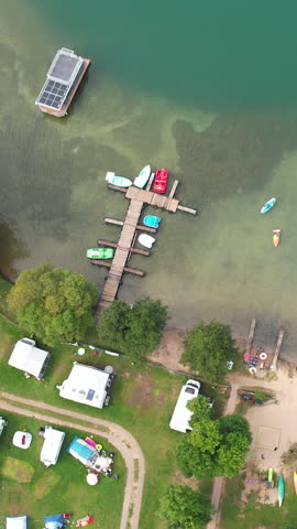 High angle cinematic view of a summer camping site by a lake in Kashubia, Poland, featuring a wooden pier, colorful boats, and parked campers.
