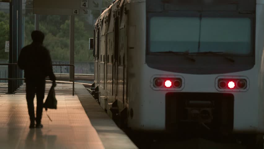 A person walks on the platform toward an arriving train during the evening time. The setting shows a station with a quiet atmosphere and urban features