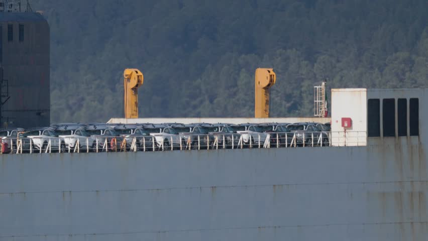 A large cargo ship is docked at a port. It is loaded with new cars lined up on the deck. Cranes stand tall next to the ship and hills rise in the background