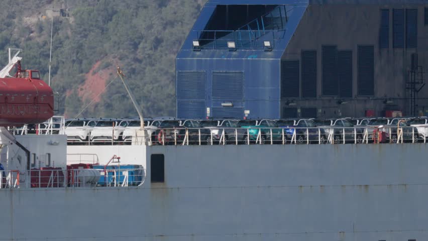 A cargo ship docks at the port carrying a collection of cars. The vehicles are lined up on the deck of the ship. The sky is clear and it is early afternoon