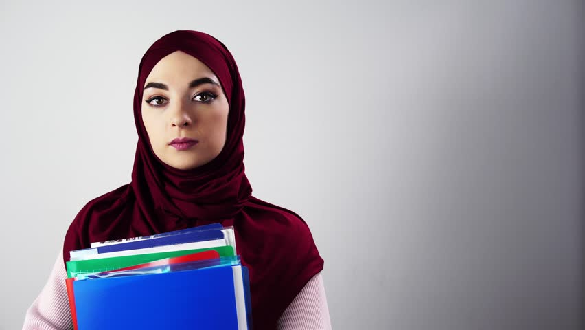 Portrait of woman in hijab holding folders and documents against neutral background, confident professional stance, clear direct gaze and neat presentation for corporate, academic, or recruitment