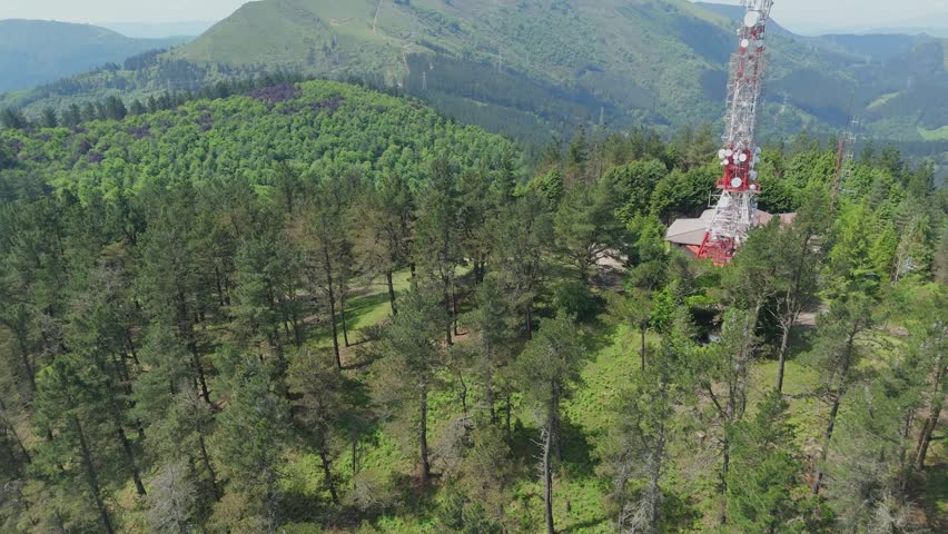 Aerial view flying backwards from a telecommunication tower on top of a mountain, revealing a vast landscape of green pine trees and dense forest under a bright sunlit day