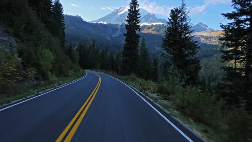 Footage captured while driving along a winding asphalt road, with double yellow lines, through a dense evergreen forest towards the snow-capped peak of Mount Rainier in Mount Rainier National Park, Wa