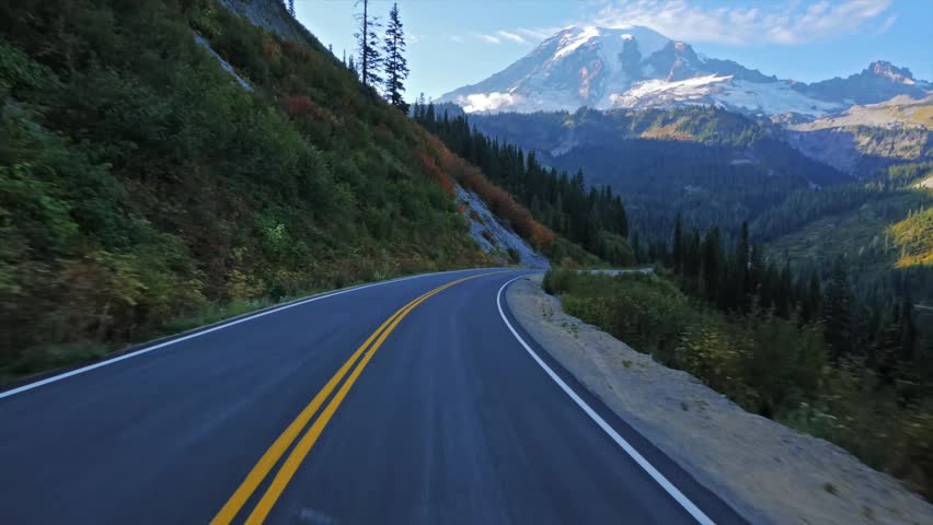 Footage of a car driving along a winding asphalt road, likely Stevens Canyon Road, near Reflection Lake in Mount Rainier National Park, Washington State, USA. The road leads towards the majestic, snow