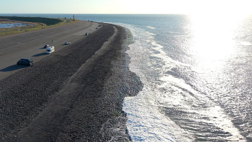 An aerial drone perspective captures the expansive stone and asphalt sea dike protecting the coast. Sun glare glints off the breaking North Sea waves, while vehicles travel along the dike road