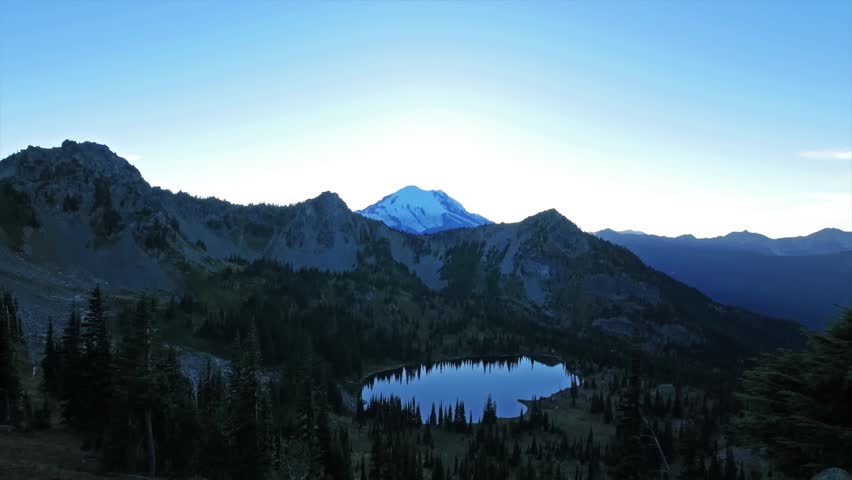 A serene, wide-angle shot captures the majestic Mount Rainier at sunrise, its snowy peak glowing blue against the clear morning sky. The view is from the Pacific Crest Trail near Sourdough Gap, overlo