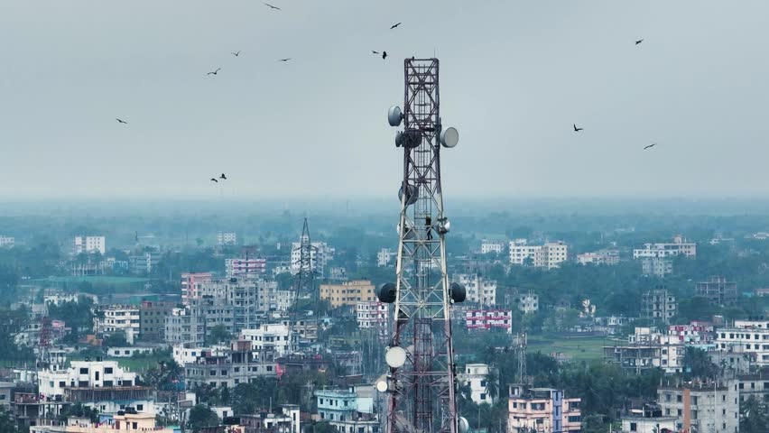 Aerial view of a telecommunications tower with a technician climbing among satellite dishes and birds flying over a dense urban landscape.