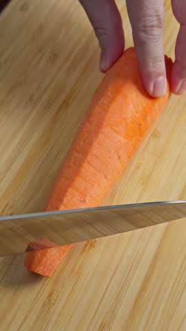 Cutting carrot on wooden cutting board for vegetable preparation. Cutting fresh carrot on a wooden cutting board and adding chopped vegetables. Home cooking preparation in kitchen.