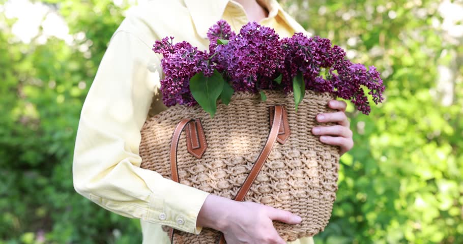 Woman in shirt holding wicker bag with beautiful lilac flowers outdoors, closeup