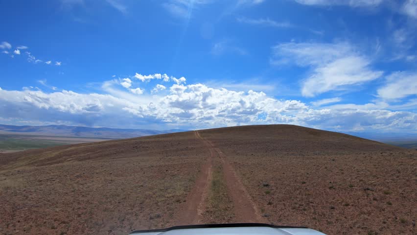 Video of a car driving along a country road under blue sky. Altai desert and semi-desert environment. The village of Kokorya is visible in the valley.