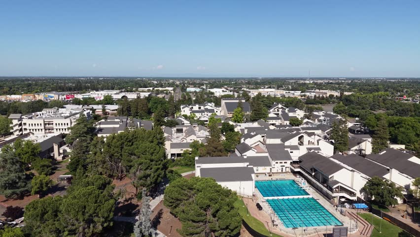 Sunny aerial perspective of the San Joaquin Delta College campus, featuring academic buildings, lush green trees, a large swimming pool, and the surrounding cityscape of Stockton, California