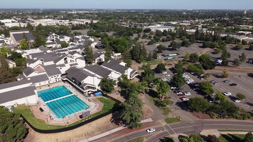 High-angle drone footage orbiting the swimming pool at the San Joaquin Delta College campus in Stockton, California, showing the surrounding buildings, parking lot, and cityscape on a sunny day