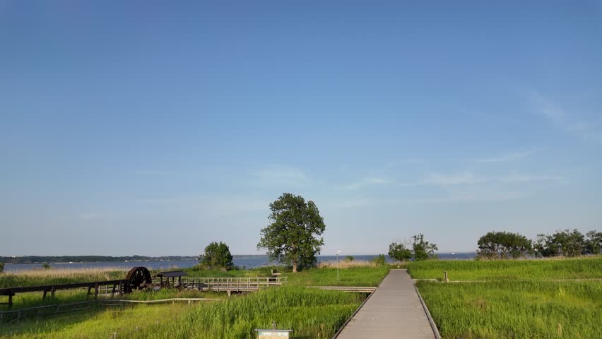 POV walking on a wooden boardwalk towards a traditional wooden water wheel in Kasumigaura Sogo Park under a clear blue sky