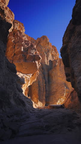 Vertical Screen: Lone hiker explores stunning Golden Canyon in Death Valley National Park, California. Towering orange and gray rock formations rise under a vivid blue sky creating dramatic shadows.