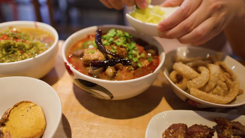 Hands squeeze fresh lime juice into a bowl of spicy Northern Thai noodle soup