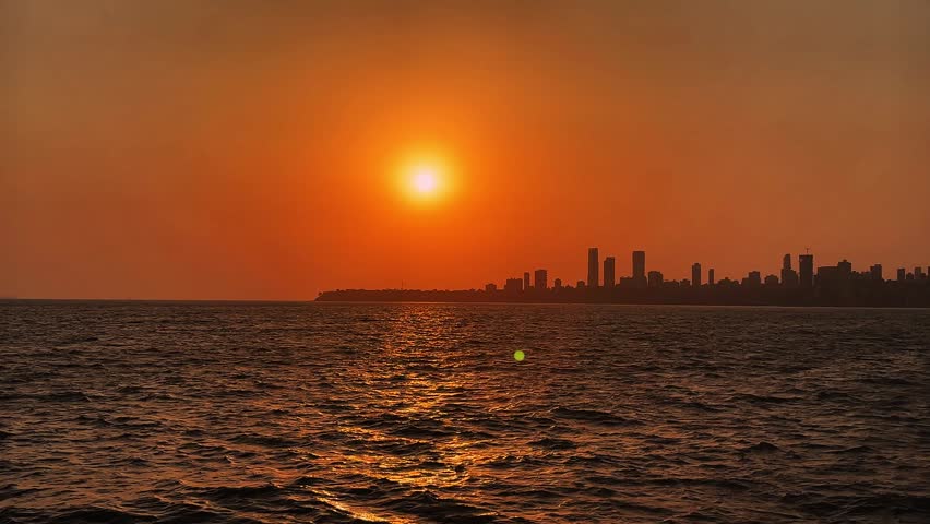 Bright orange sunset over coastal city skyline with shimmering sea water and evening light.
