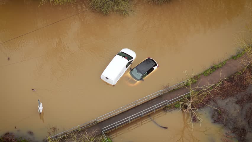 Cars submerged in floodwater during heavy rain season across Britain. Flash flooding covering country road and trapping vehicles near small pedestrian bridge. Torrential rainfall hitting England and