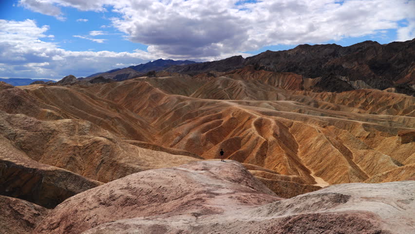 Solitary figure stands atop colorful badlands at Zabriskie Point in Death Valley National Park. Golden and ochre sedimentary ridges stretch toward distant mountains under dramatic cloudy skies.