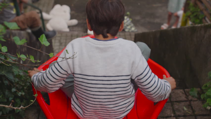 Boy slides down the stairs in a red sled, leaning forward with eager determination as siblings wait below, forming a lively shared outdoor play moment