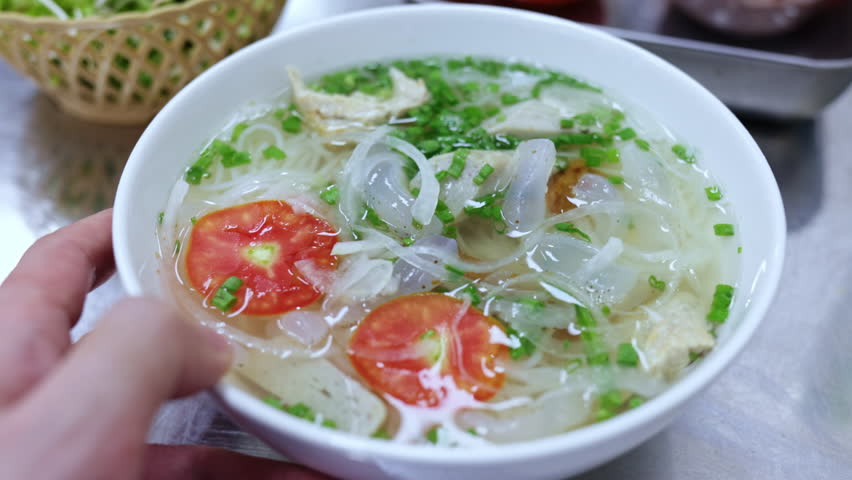 Person holds a white bowl of traditional vietnamese bun ca, a steaming fish noodle soup with jellyfish, tomatoes, onions, fishcake, herbs, and clear broth, ready for a delicious meal