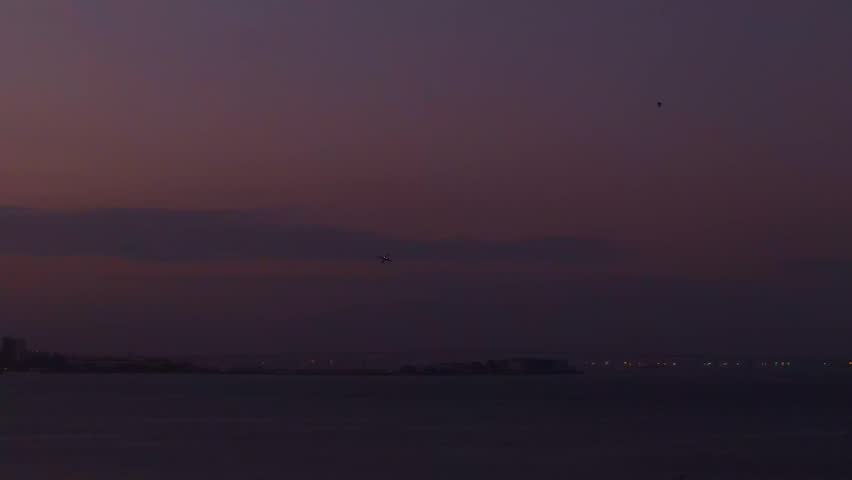 An airplane taking off from Santos Dumont Airport at dusk, with the Rio-Niteroi Bridge in the background under a hazy sky in Rio de Janeiro, Brazil.