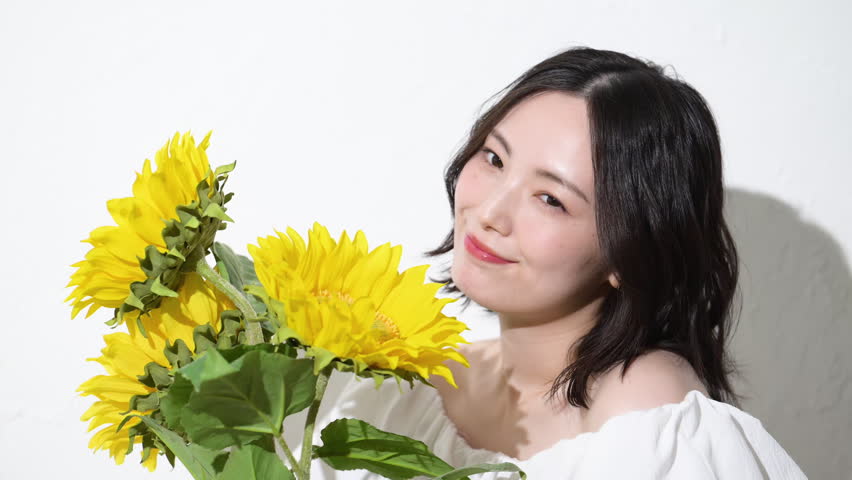 A Japanese woman smiling while holding a sunflower. A video evoking images of summer beauty and a cheerful lifestyle. She looks directly at the camera.