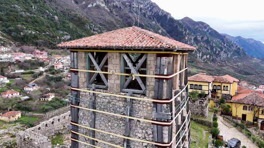 Aerial camera circles counterclockwise around the historic stone Clock Tower under restoration within Kruja Castle, Albania, framing the rugged mountain landscape in the background