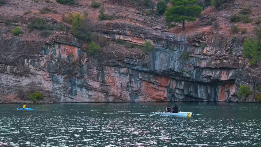 Cinematic wide view of two rowers moving across a calm lake beneath rocky cliffs. Peaceful outdoor sports scene with freshwater, natural landscape, motion and open water.