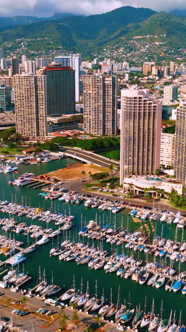 Aerial view of busy marina filled with docked sailboats and yachts, fronting Honolulu high-rise towers with green mountain backdrop. Vertical video.