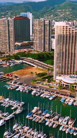 Aerial view of Honolulu marina with sailboats and motorboats docked along piers, high-rise hotel towers and lush green mountains behind. Vertical video.