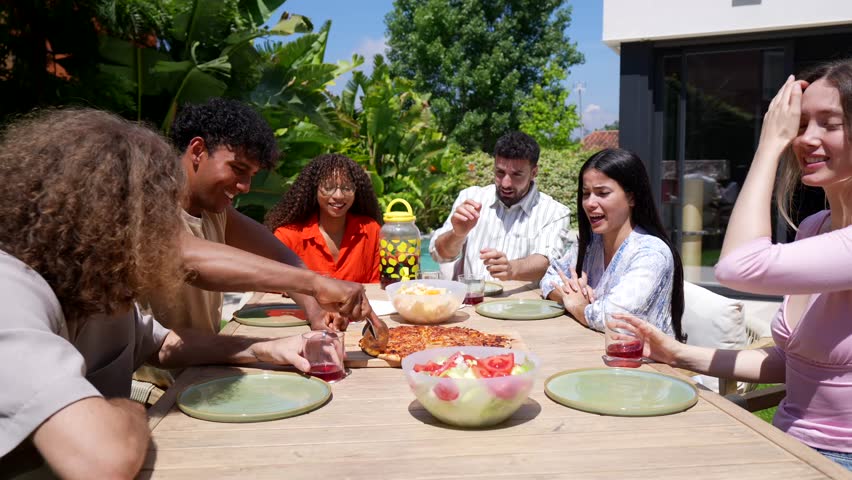 Happy group of diverse young friends enjoying an outdoor lunch together in a sunny backyard, laughing and sharing slices of freshly cut pizza during a cheerful summer gathering at home