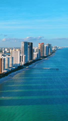 Wide aerial view of long Miami Beach sandy shoreline with blue-green ocean, high-rise hotels, fishing pier and clear blue sky. Vertical video.