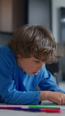 Focused young boy in a blue shirt lying at a table and drawing with colorful pencils, concentrating on a creative activity indoors