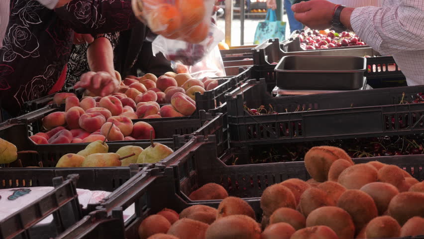 Shoppers Selecting Fresh Fruit At Busy Outdoor Farmers Market Stall. Shoppers reach for peaches and oranges at a busy outdoor farmers market stall, browsing fresh produce crates while vendors weigh and pack fruit. Casual market scene conveys community, summer and seasonal abundance.